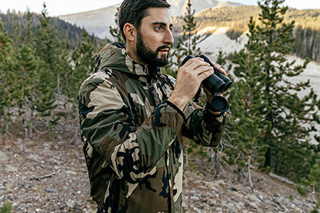 Hiker using binoculars on a mountain side while wearing the Ultra Lochi Jacket in Woodland.