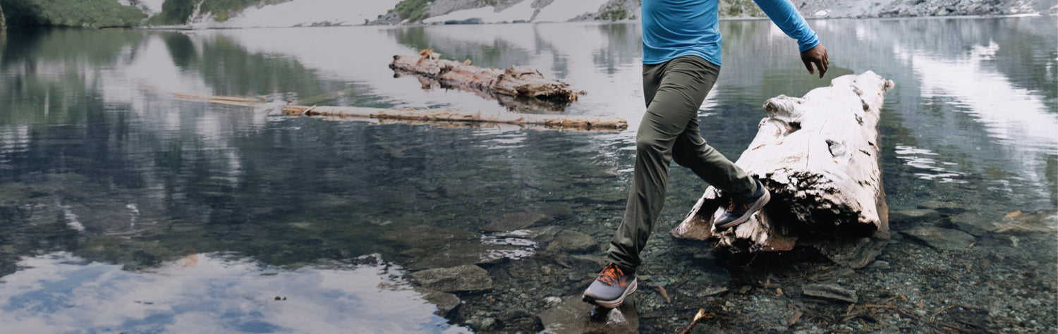 Man stepping from a log in a lake back to the shore wearing the Ascent-Glide pant in Olive Green