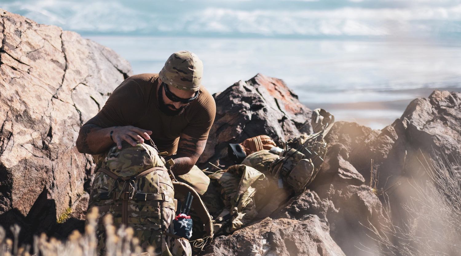 Person in camouflage gear sitting on rocks by a body of water