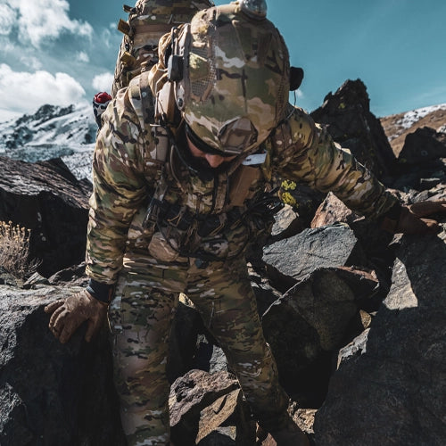 Person in camouflage gear climbing rocky terrain with mountains in the background