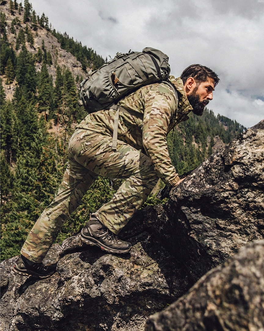 A man in the L4 Back Country LT Jacket and pants with a large olive green backpack is climbing a rocky mountain path with pine trees and cloudy skies in the background.