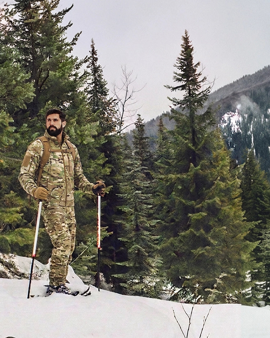 A man wearing the L5 Back Country Jacket and Pants stands in the snow holding ski poles, with a forest and mountains in the background.