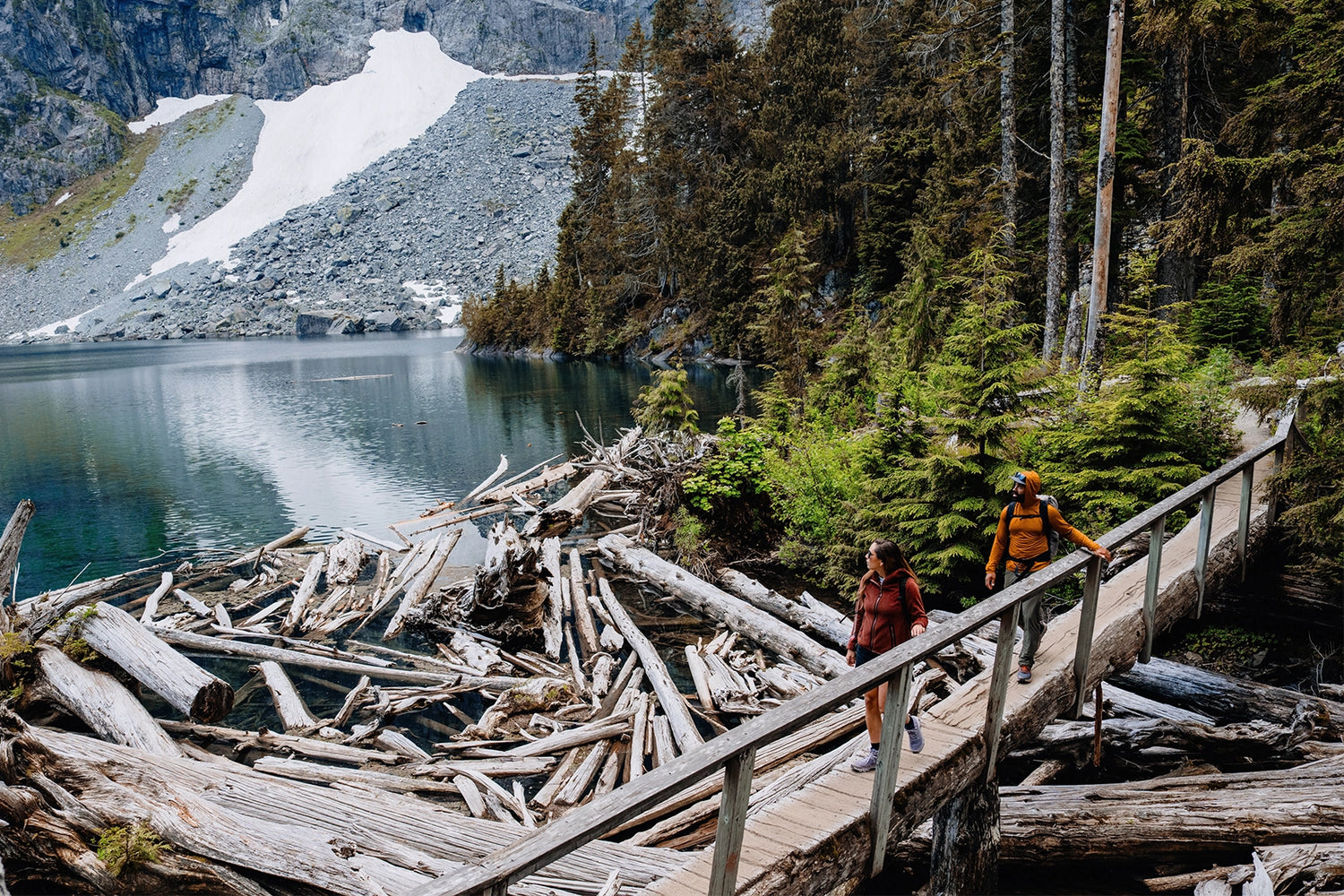 Two hikers crossing a bridge over an alpine lake. The lead hiker is wearing an Women's Alpha Aura Jacket made by Beyond Clothing and second hiker is wearing the Ascent-Glide Pant and the Mens Hooded Geo-T.