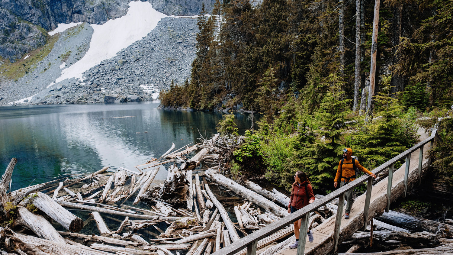 Two hikers crossing a bridge over an alpine lake. The lead hiker is wearing an Women's Alpha Aura Jacket made by Beyond Clothing and second hiker is wearing the Ascent-Glide Pant and the Mens Hooded Geo-T.