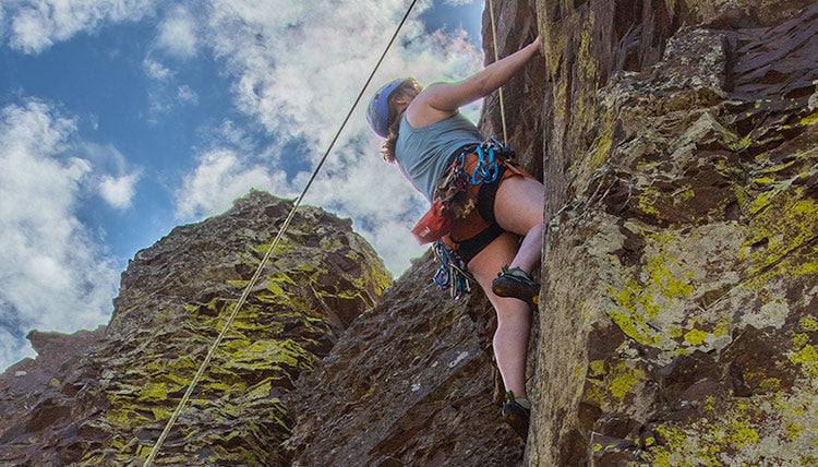 Woman wearing the Big Sky Blue Feathers Tech-Tank and the Desert Clay Olallie Loop Shorts while rock climbing with a partially cloudy sky in the background.