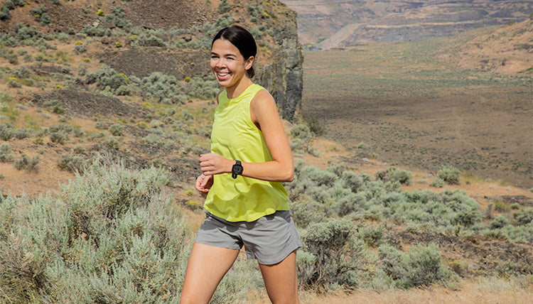 Woman trail running in a high desert environment while wearing the Light Lime colored Feathers Tech-Tank and the Moonstone colored OlallieLoop Shorts. Running towards the camera with a smile.