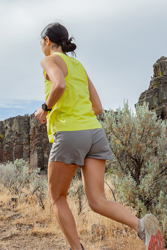 Woman trail running in a high desert environment while wearing the Light Lime colored Feathers Tech-Tank and the Moonstone colored OlallieLoop Shorts. Running away from the camera.. 