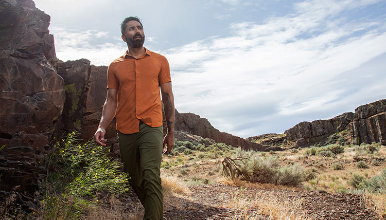 Man walking in a high desert landscape with rock formations in the background while looking into the distance. He is wearing the Desert Clay FirstSun Shirt and the Evergreen TarnGood Pant.