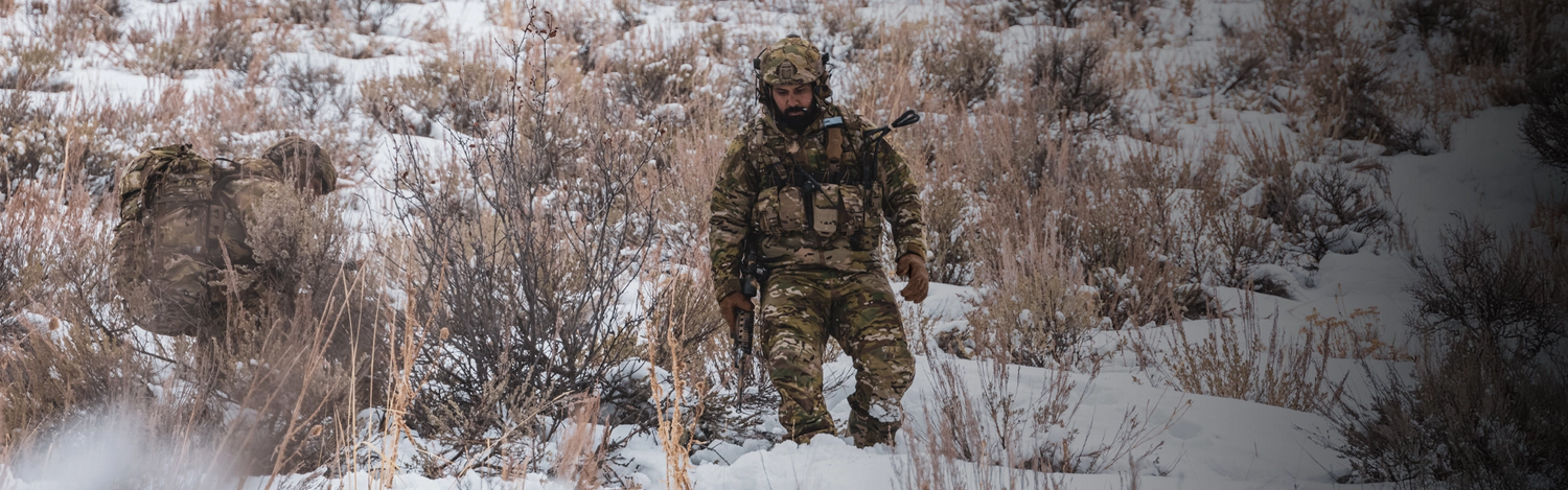 Person in camouflage gear walking through a snowy landscape with dry grass.