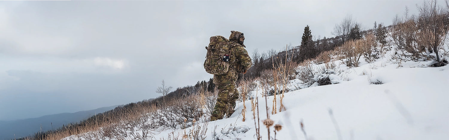 Person in camouflage gear with a backpack walking on a snow-covered landscape.
