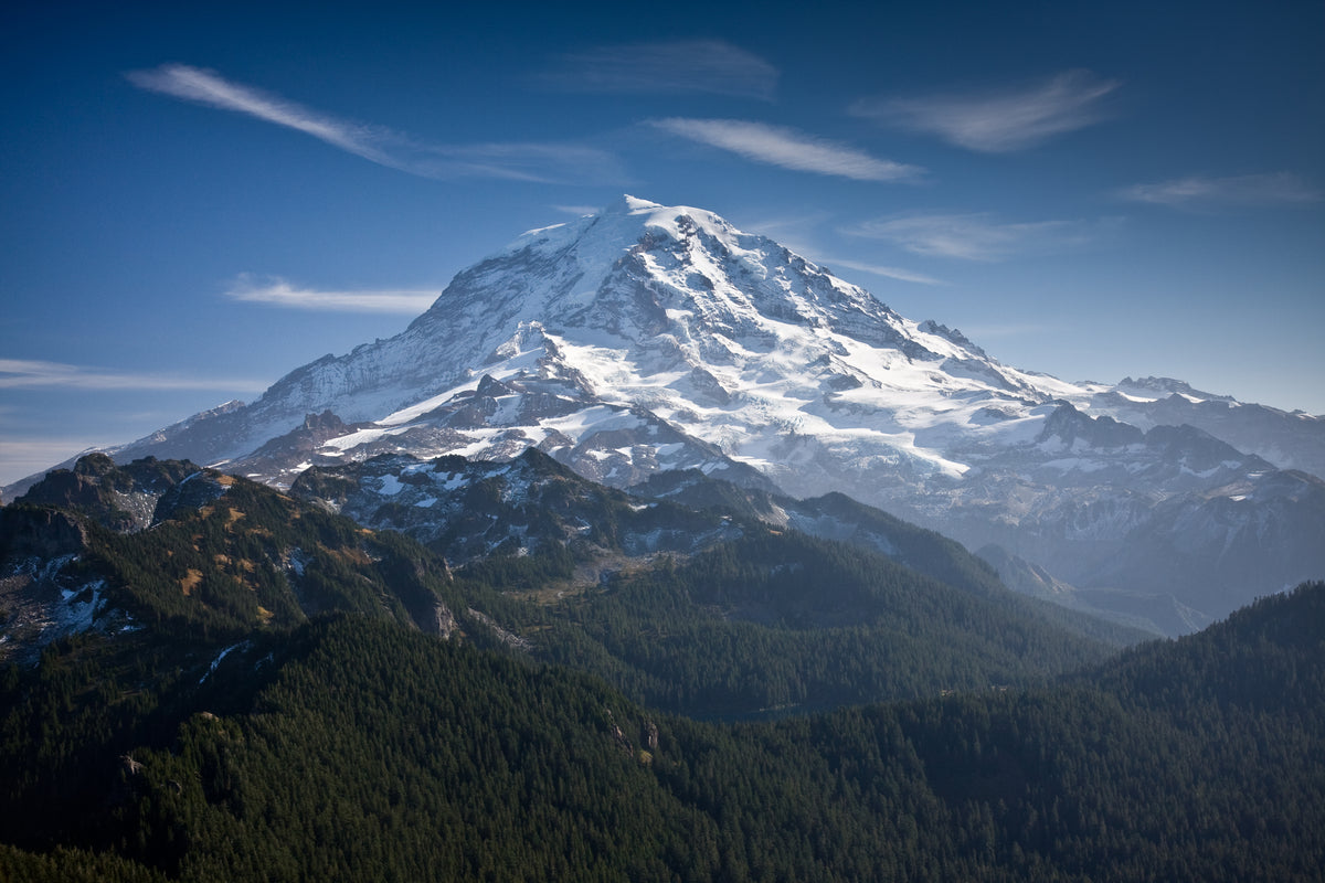 Mount Rainier a majestic snow-covered mountain peak rising above dark green forested hills under a clear blue sky with wispy clouds.
