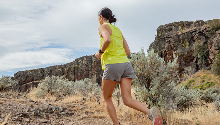 Woman trail running in a high desert environment while wearing the Light Lime colored Feathers Tech-Tank and the Moonstone colored OlallieLoop Shorts. Running towards the camera with a smile.