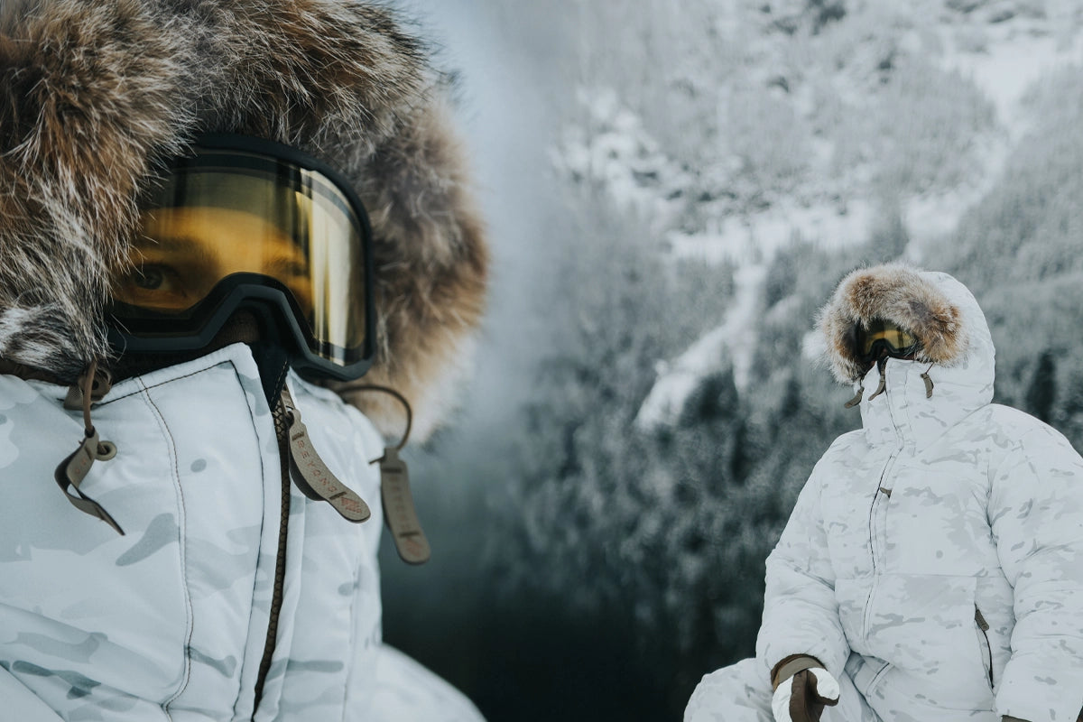 Two people in winter clothing with fur hoods and goggles against a snowy mountain backdrop.
