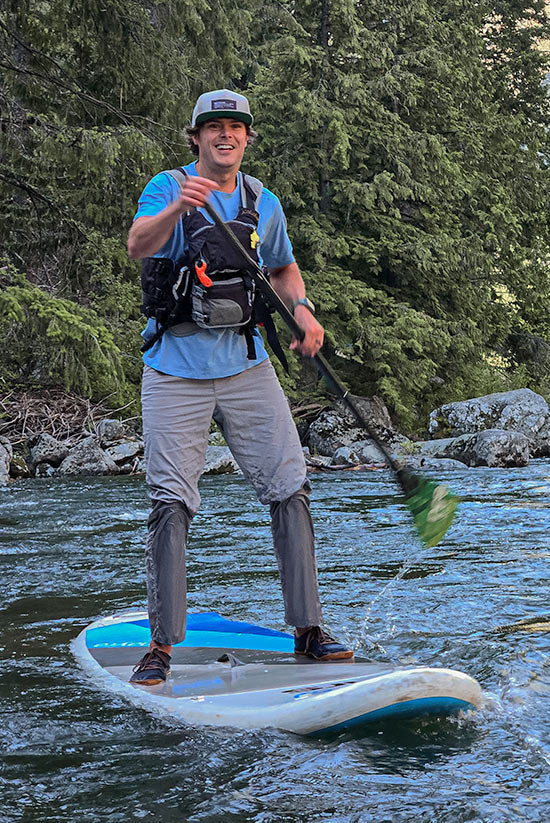 Man paddle boarding in a forested river while wearing the Moonstone TarnGood pants and the Big Sky Blue SolarSwift Tech-T. Smiling looking towards the camera. 