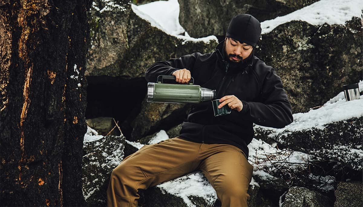 Man sitting on a snowy mountain side pouring his thermos into a mug while on a break during a hike.