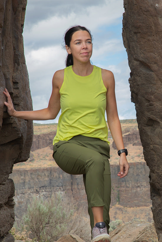 Woman wearing the Light lime Feathers Tech-Tank and the Evergreen Tinkham pant while standing between two rock formations. 